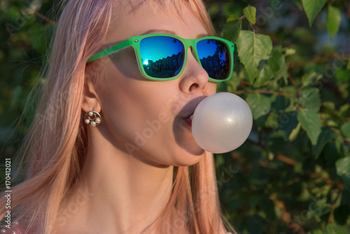 Fotografie Young girl in green glasses with long hair puffs bubble of chewing gum in summer