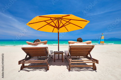 Fototapeta Naklejka Na Ścianę i Meble -  Family couple on tropical beach on deck chairs under orange umbrella