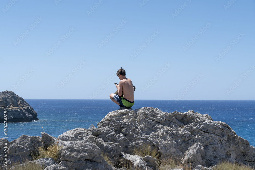 young man crouching on a rock with mobile phone young man crouching on a rock with mobile phone