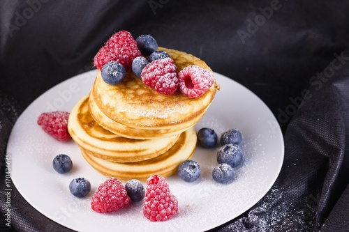 Stack of pancakes with raspberries and blueberries  coated with powdered sugar on white plate and black background