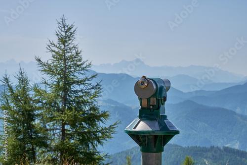 Tourist telescope pointing at misty covered mountains from the top of the Panoramastrasse on Mt. Goldeck with a pine tree standing in the foreground photo