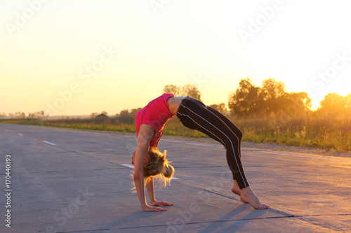 Yoga professional performing pose and handstand. Peaceful healthy young girl doing power pose on rural backstage, road on sunset. Slim woman doing yoga over orange sun.  photo