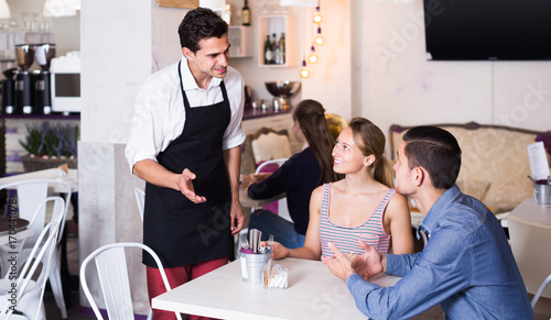 Portrait of young cafe waiter standing at table and talking to guests