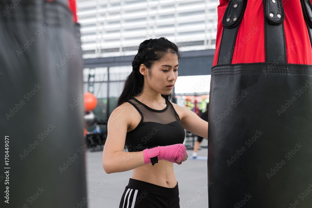 young woman execute exercise in fitness center. female athlete hitting and punching sandbag with rowing machine in boxing gym. sporty girl working out in health club. martial arts, muay thai concept
