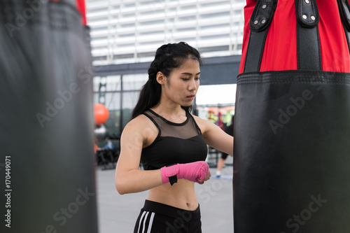young woman execute exercise in fitness center. female athlete hitting and punching sandbag with rowing machine in boxing gym. sporty girl working out in health club. martial arts, muay thai concept © 88studio