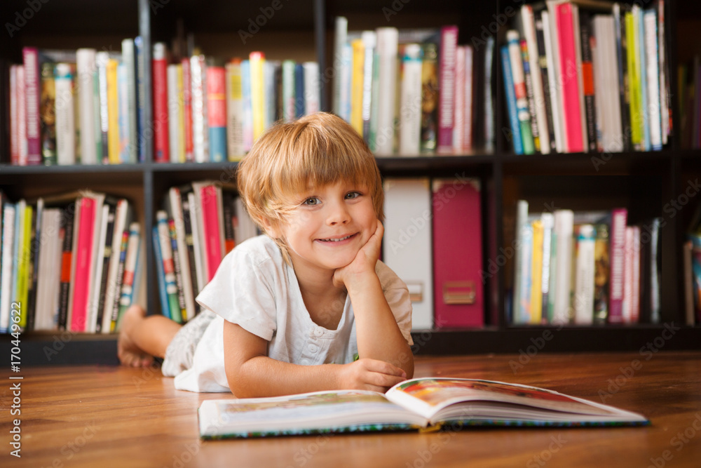 Cute little boy is reading book sitting near bookshelf at home Cute little boy is reading book sitting near bookshelf at home