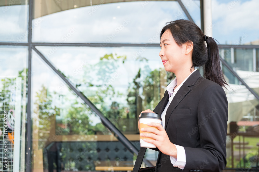happy businesswoman holding coffee outside office building. beautiful young asian woman with tea go to work outdoors. female adult with disposable paper cup walking in business area