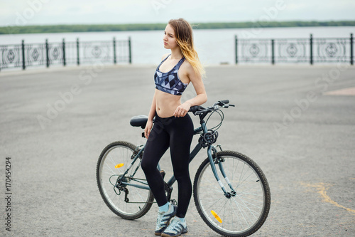 Young seductive fitness woman in sport wear posing outdoor on the bicycle at river bank
