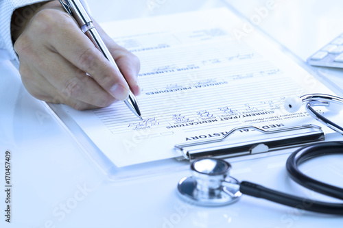Closing up doctor’s hand writing medical history record on clear transparent clipboard with black stethoscope on glossy desktop. Presented in light blue-white balance.