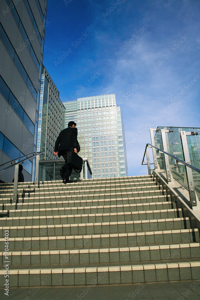 A low angle photo of a businessman running up a set of stairs towards blue sky in Gotanda, Japan