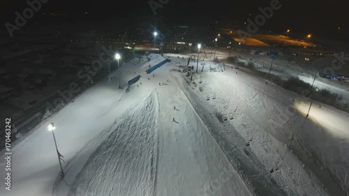 Aerial view: Skiers and snowboarders going down the slope in winter night. Skiers and snowboarders enjoying on slopes of ski resort in winter season. 4K video, aerial footage.