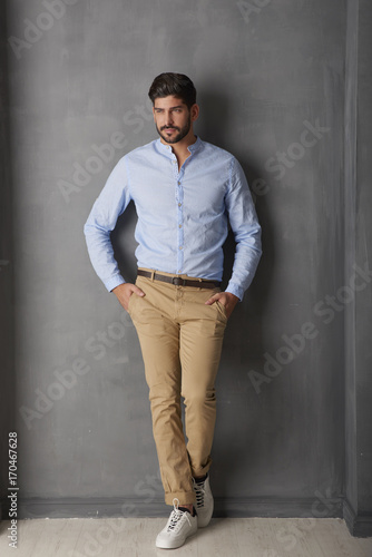 Relaxed young man looking at camera and smiling while standing at grey wall in the studio.