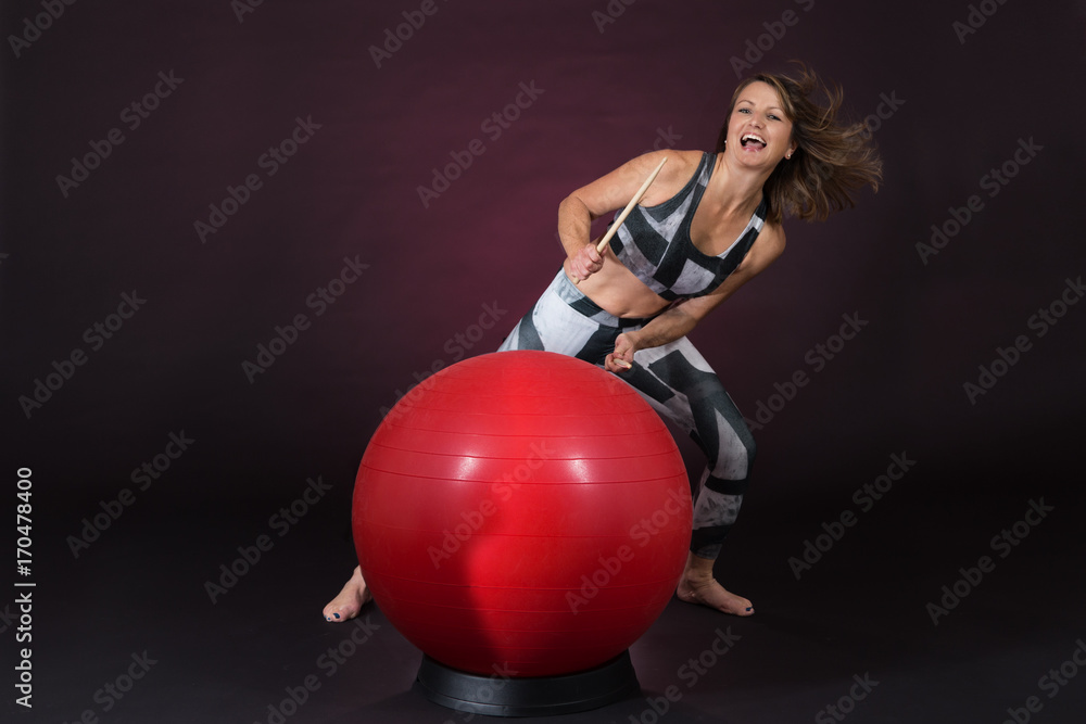 Reifere Frau im Sport Outfit trommelt mit Drum Sticks auf einem roten Gymnastikball vor einem dunklen Hintergrund Reifere Frau im Sport Outfit trommelt mit Drum Sticks auf einem roten Gymnastikball vor einem dunklen Hintergrund