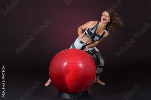 Reifere Frau im Sport Outfit trommelt mit Drum Sticks auf einem roten Gymnastikball vor einem dunklen Hintergrund