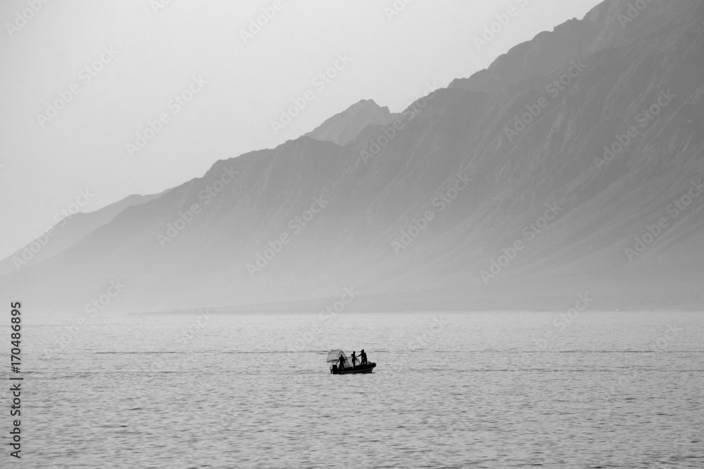Picturing the seascape in the morning with the background is Rocky mountain. A silhouette of fishermen on the boat while fishing. The picture is in black and white. Picturing the seascape in the morning with the background is Rocky mountain. A silhouette of fishermen on the boat while fishing. The picture is in black and white.
