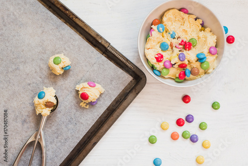Cookie dough with colorful chocolate candies in bowl next to dough scooper and scoops of cookie dough on baking sheet