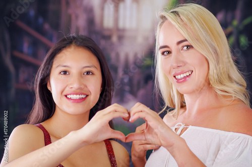 Two young women blonde and latin girl smiling and breaking racism idiosyncrasy from a american person and foreign people, both doing a heart sign with teir hands, racism, violence or discrimination photo