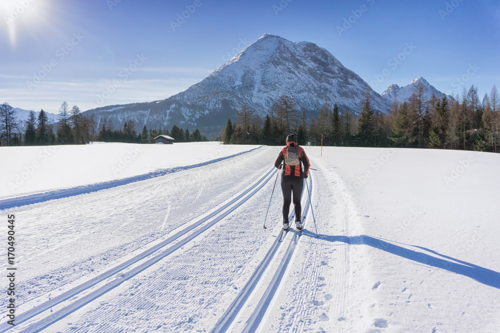 Cross-country Skie in red jacket runs on groomed ski track in sunny winter day. Winter mountain landscape: Tirol, Alps, Austria.