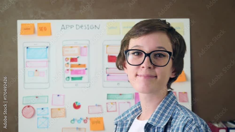 Young UX designer looking at the whiteboard with design layout then turns around smiling and looking at the camera Young UX designer looking at the whiteboard with design layout then turns around smiling and looking at the camera
