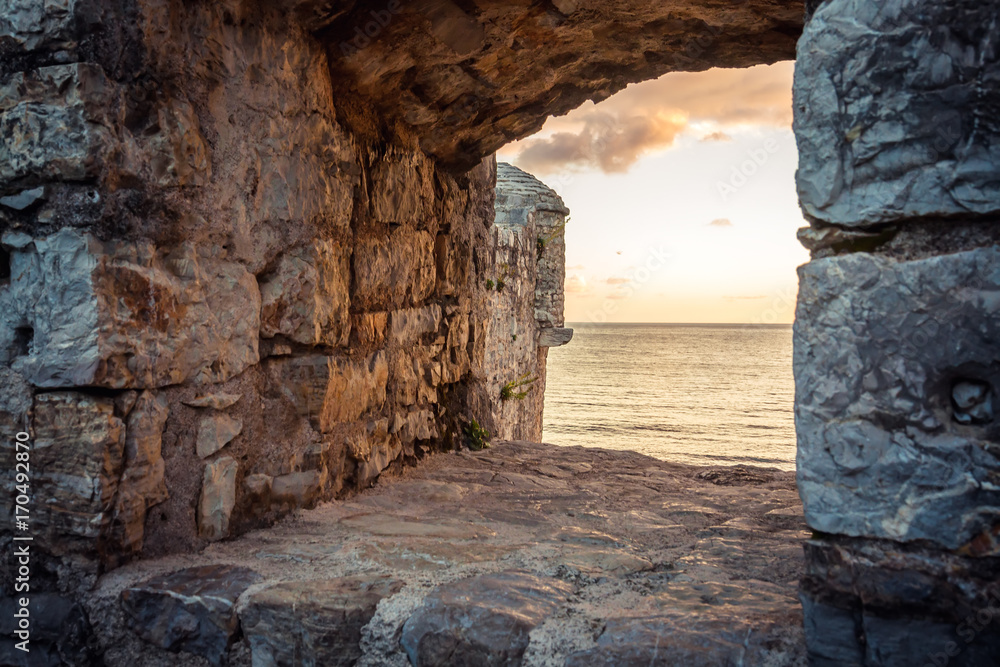 old ruins background with scenic sunset over sea through ancient castle window with dramatic sky and perspective view with effect of light at the end of tunnel as travel background