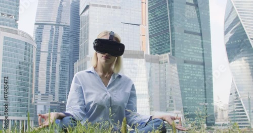 A woman in a blue shirt and VR glasses is sitting in a lotus pose. Skyscrapers are on background