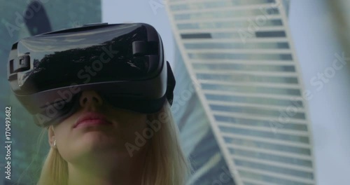 A woman in a VR glasses is sitting in a lotus pose. Skyscrapers are on background