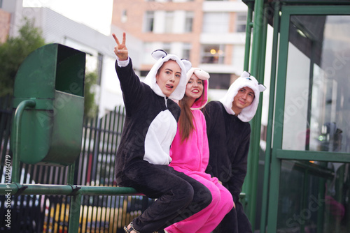 Close up of a happy group of friends, waiting the public transportation and wearing different costumes, one woman wearing a pink unicorn costume, other woman a panda costume and the man wearing a cat
