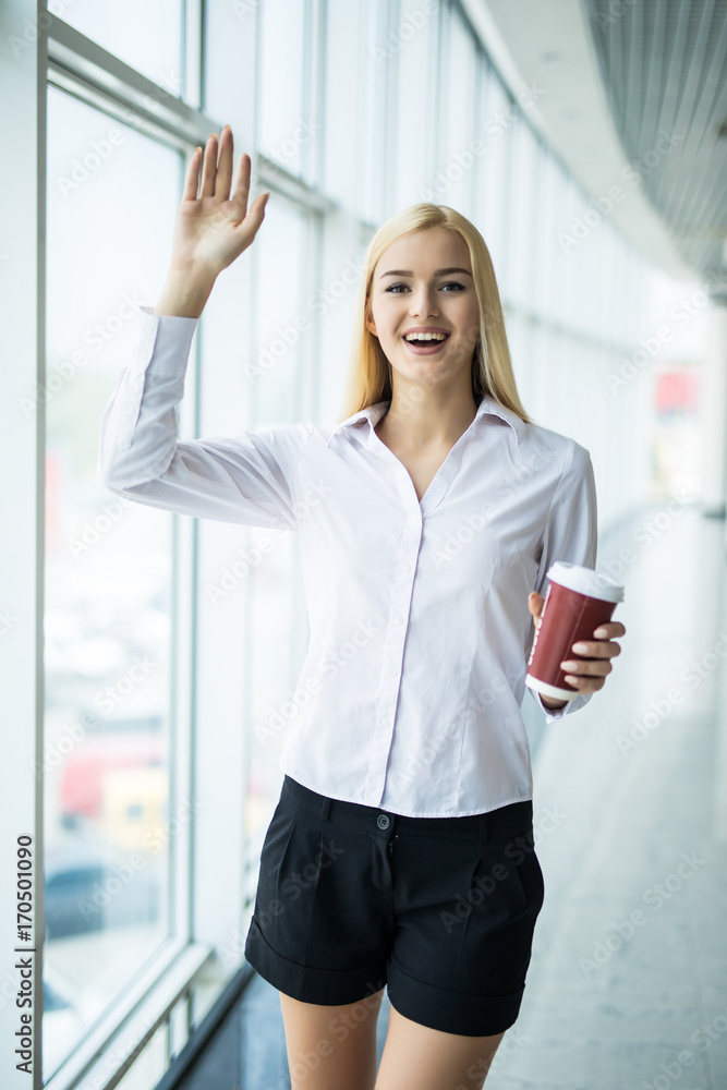 Businesswoman welcomes with cup of coffee in other hand near big panoramic window in the office Businesswoman welcomes with cup of coffee in other hand near big panoramic window in the office