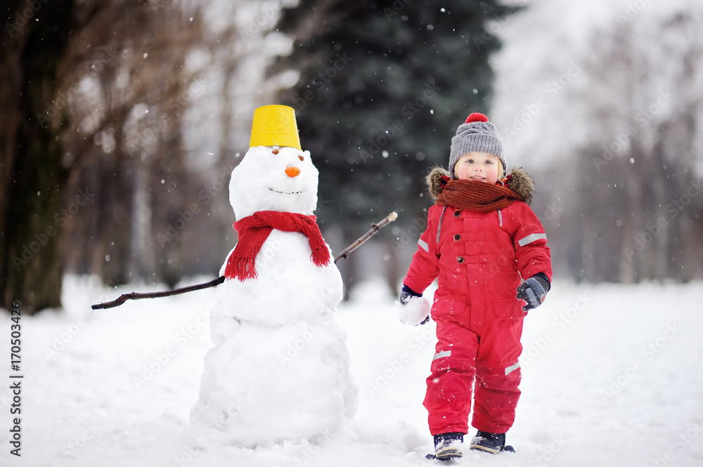 Little boy in red winter clothes having fun with snowman in winter park Little boy in red winter clothes having fun with snowman in winter park