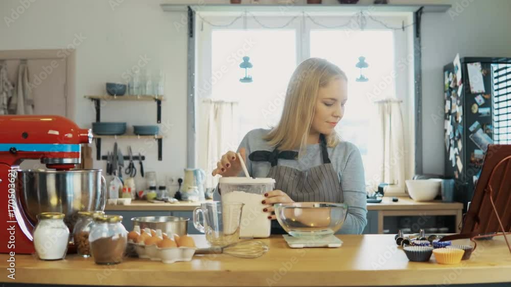 Young blonde woman using the kitchen scales to weighing the flour. Beautiful female baking the cupcakes on the kitchen. Young blonde woman using the kitchen scales to weighing the flour. Beautiful female baking the cupcakes on the kitchen.