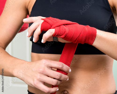 Young active woman getting prepared for exercises wrapping her hands with red bandage tape in homemade gym, fitness