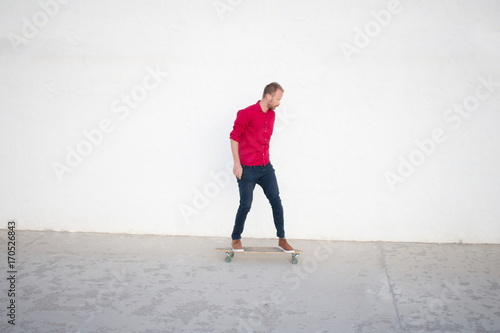 Young bearded man riding on skateboard, hipster with longboard in red shirt and blue jeans urban background 