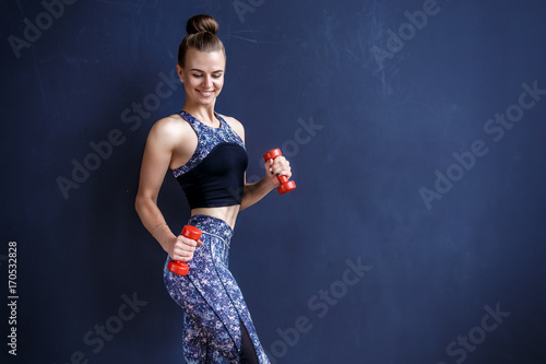 Beautiful fitness model is posing in front of the dark wall in a dark training suit