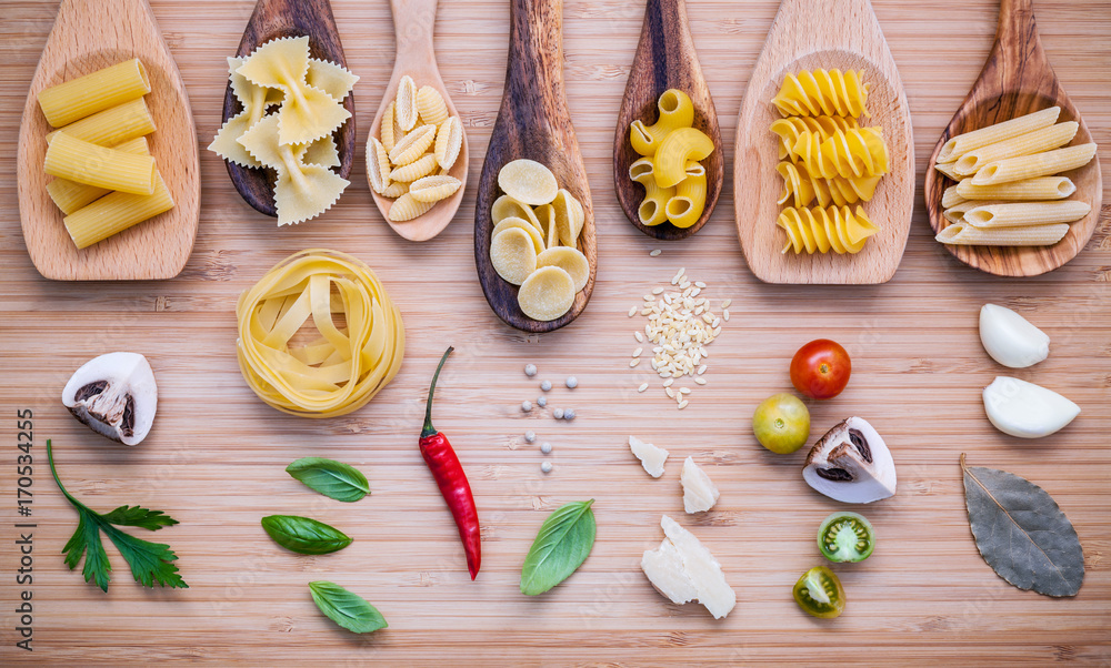 Italian food concept .Various kind of pasta in wooden spoons with ingredients sweet basil ,tomato ,garlic ,parsley ,bay leaves ,pepper ,champignon and parmesan cheese on bamboo cutting board flat lay. Italian food concept .Various kind of pasta in wooden spoons with ingredients sweet basil ,tomato ,garlic ,parsley ,bay leaves ,pepper ,champignon and parmesan cheese on bamboo cutting board flat lay.