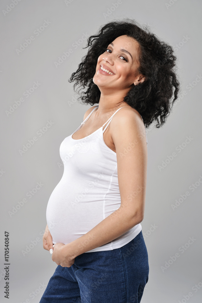 Pregnant woman smiling looking at camera with hands on tummy, over grey background Pregnant woman smiling looking at camera with hands on tummy, over grey background