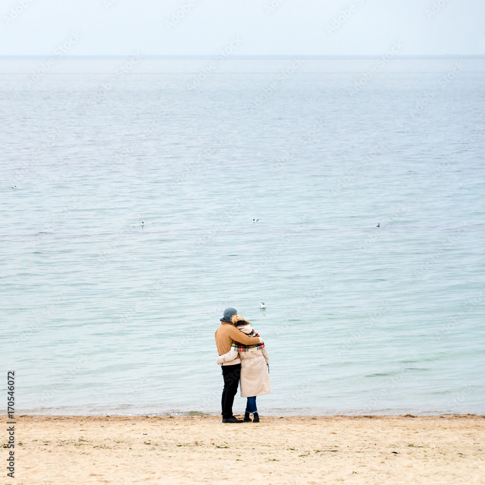 Happy loving couple embracing wearing warm clothes standing on ocean coast. Cold season, sea shore, empty beach in autumn or winter.