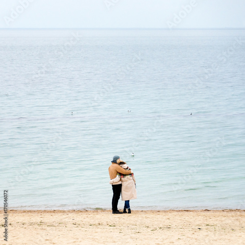 Happy loving couple embracing wearing warm clothes standing on ocean coast. Cold season, sea shore, empty beach in autumn or winter.