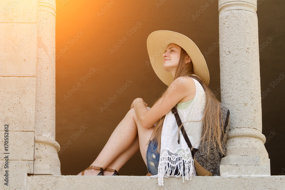 Girl in a hat and with a backpack is sitting near the column. Angle from below