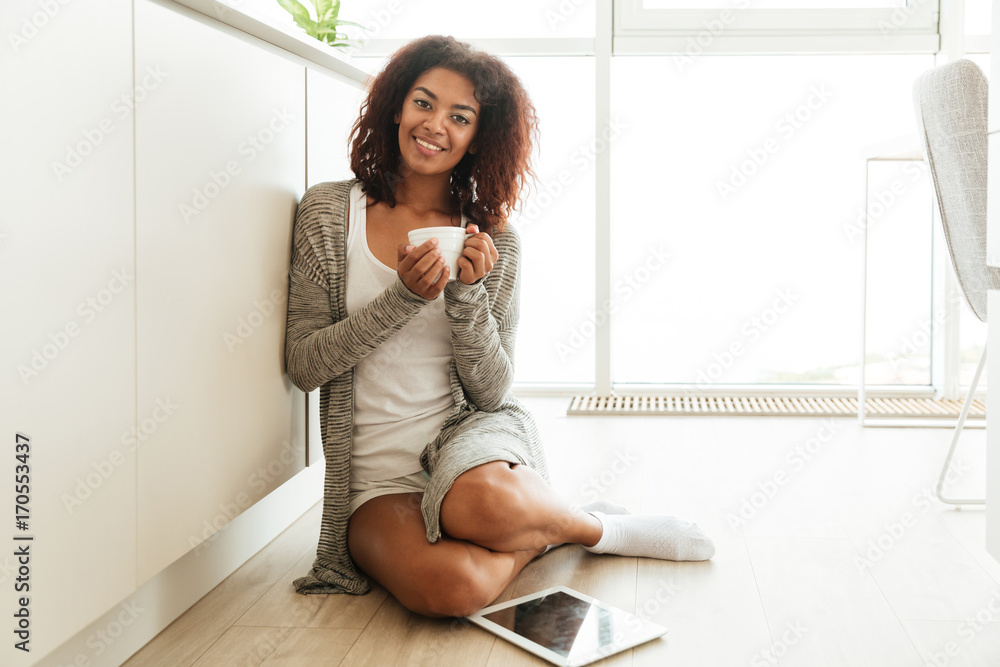 Cheerful woman with cup of tea and tablet sitting on floor