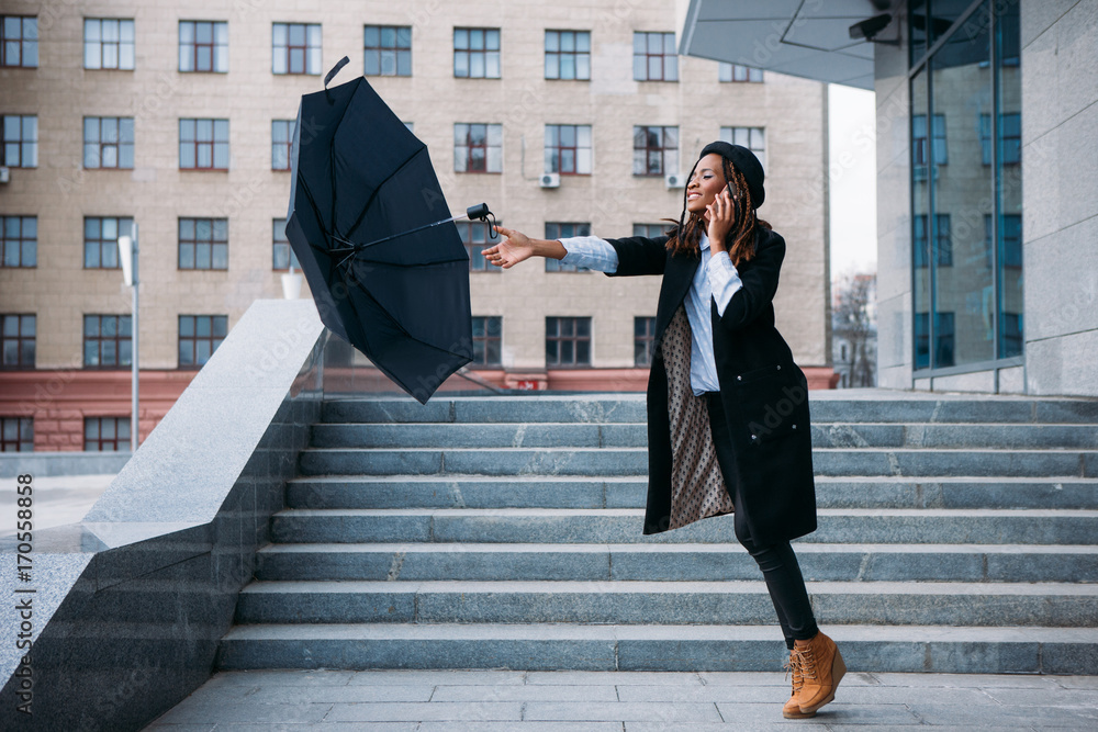 Stormy weather. Black girl catch flying umbrella speaking on mobile phone on street, airiness concept Stock-foto | Adobe Stock