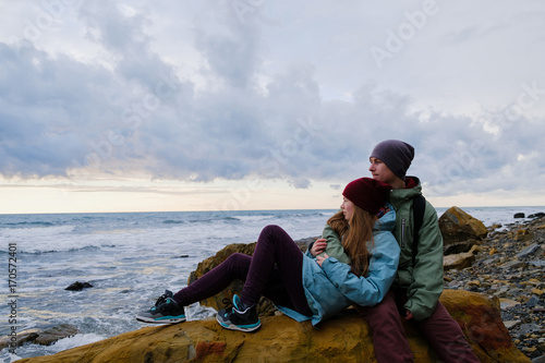 young couple sitting on rocks on the shore, hugging and looking at sea