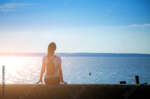 Girl sits on the stone embankment of coast sea and looks at the sun reflects on the surface of the water. In the backlight sunbeam light. Concept theme: travel, meditation, contemplation, relax, quiet