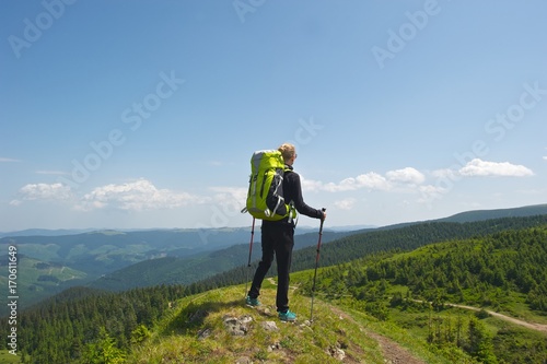 A young woman standing on top of a hill, preparing herself for the upcoming adventure. Beautiful view of the sky, trees and mountains. The woman is wearing black clothes and bright green backback. 