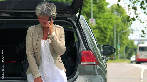 Middle aged woman sits in the trunk (car stands on the verge of road) and calls with smartphone - street with passing cars in the background