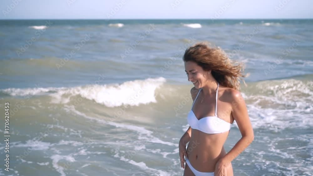 Young beautiful sexy tanned girl in bikini with curly hair sitting on the beach at sunset by the sea. She smiled enigmatically, stroked herself and. Slow motion. 120fps Young beautiful sexy tanned girl in bikini with curly hair sitting on the beach at sunset by the sea. She smiled enigmatically, stroked herself and. Slow motion. 120fps