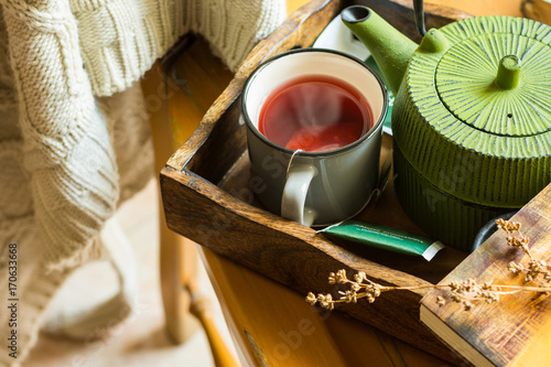 Mug with hot red fruit tea, green pot, book on tray, knitted sweater hanging over wooden chair, cozy atmosphere, autumn, fall