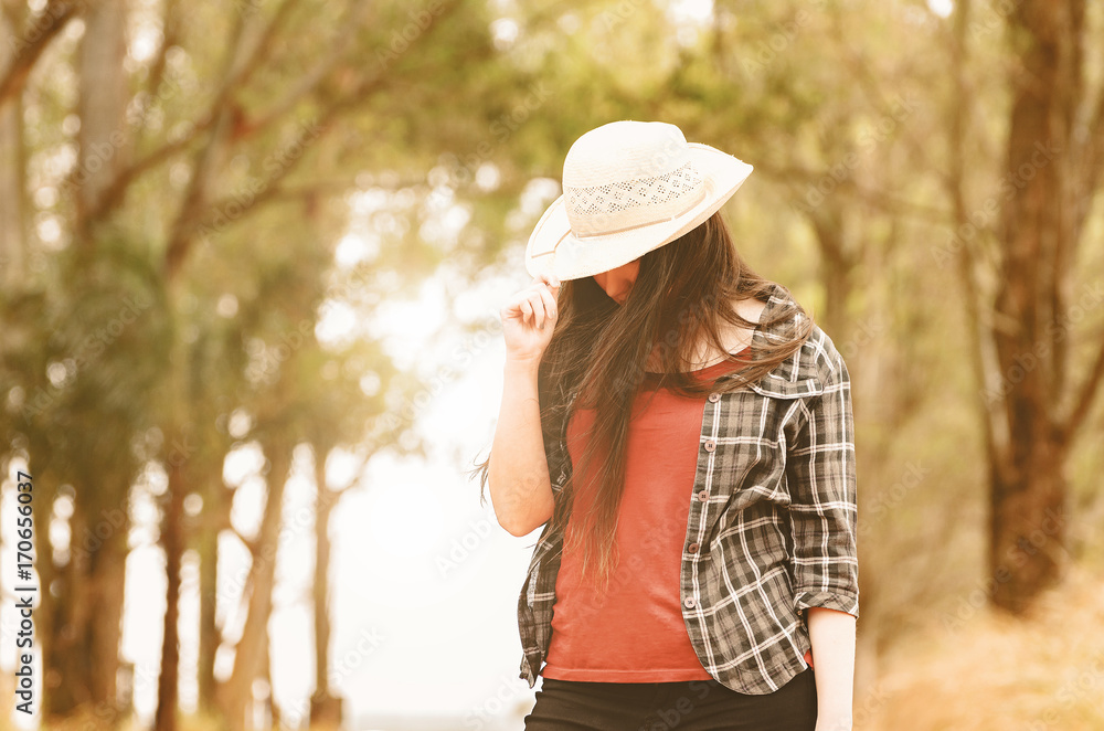 Woman hiding her face with a hat. Cowgirl on a farm wearing a striped T-shirt, in the background trees in a rural scene.
