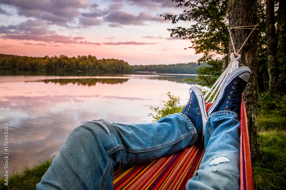 Summer camping by the lake. Young man wearing jeans and sneakers relaxing in the hammock at sunset.  - obrazy, fototapety, plakaty 