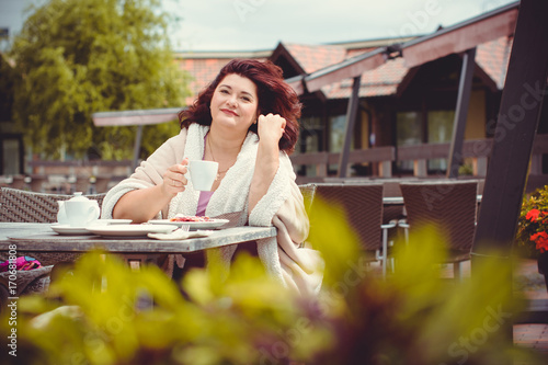 A mature woman sits in a cafe on the terrace in the autumn day, wrapped up in a warm cozy blanket and looks forward to the arrival of friends. Life of women after 40 years, their problems and joys