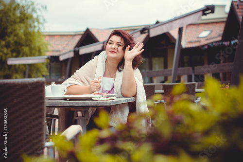 A mature woman sits in a cafe on the terrace in the autumn day, wrapped up in a warm cozy blanket and looks forward to the arrival of friends. Life of women after 40 years, their problems and joys